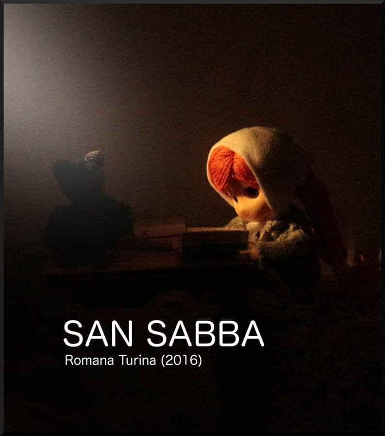 small-scale model character with a head scarf reading a book. The words: San Sabba, Romana Turina (2016) underneath