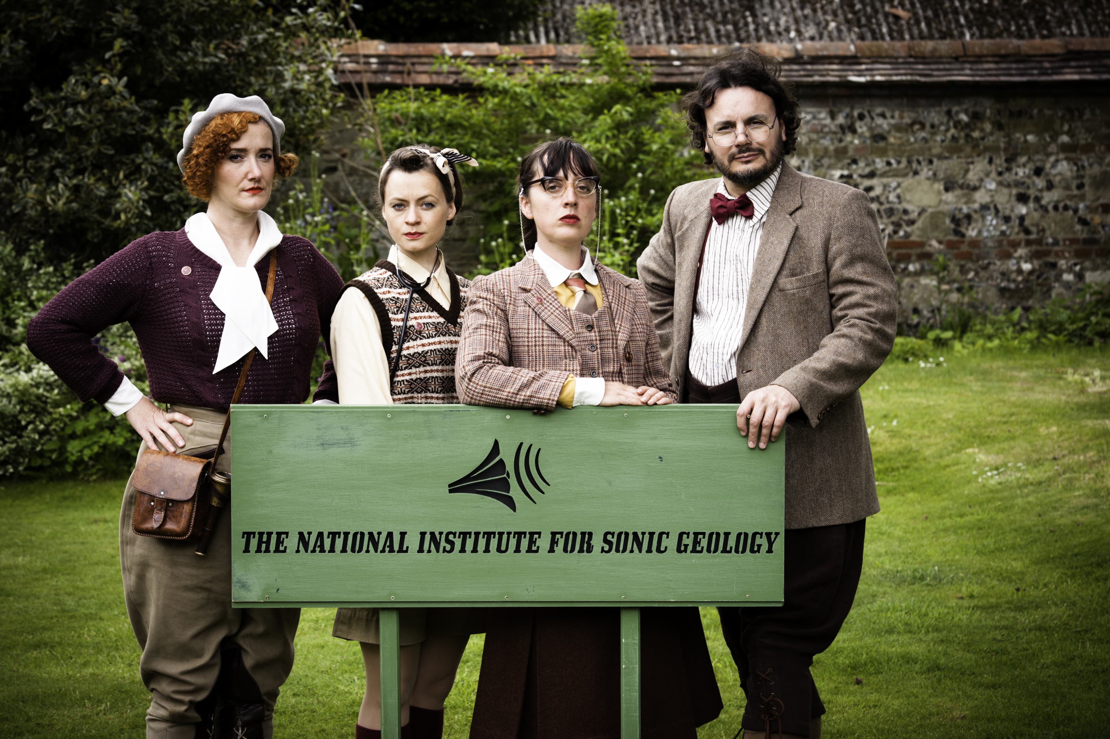 An image if 4 period outside in a park wearing brown and tweed outfits standing in from of a sign that reads The National Institute for Sonic Geology