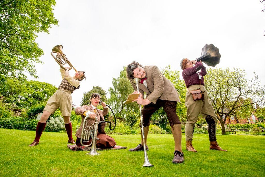 An image of 4 period outside in a park wearing brown and tweed outfits holding Ear Trumpets made from old brass instruments