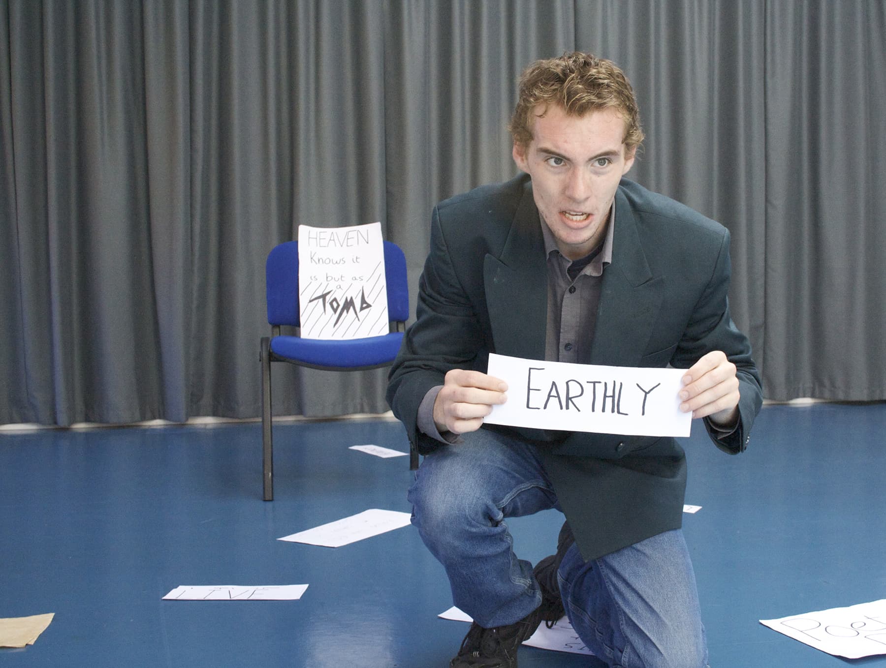 1 Acting student performing on stage in front of a curtain, holdin a sign that says 'EARTHLY'