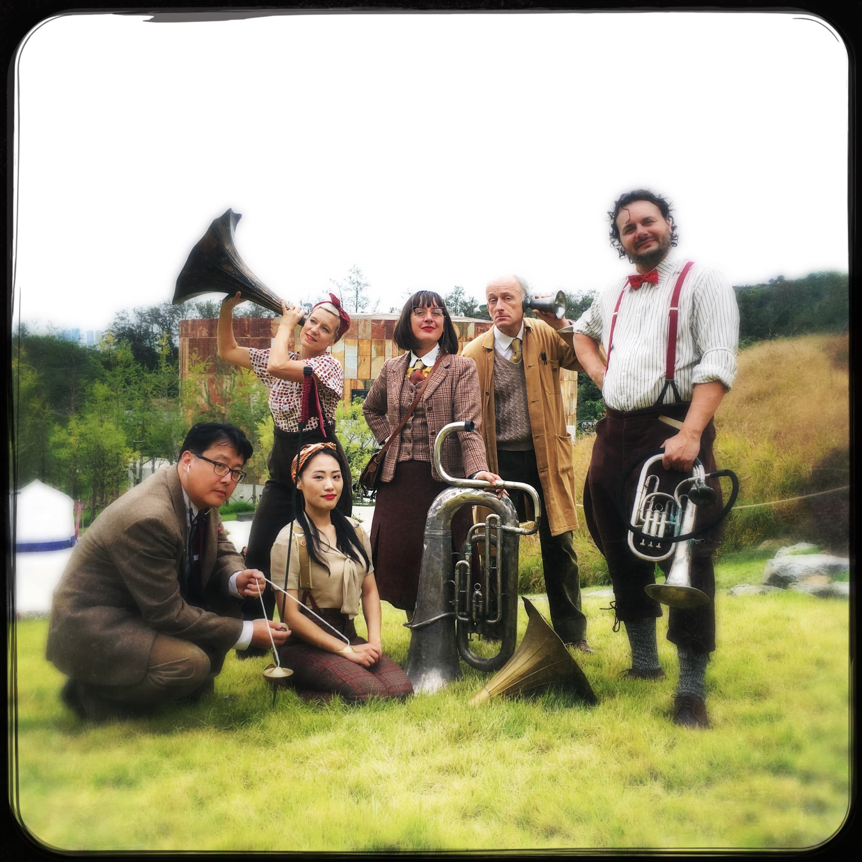 An image if 4 period outside in a park wearing brown and tweed outfits holding Ear Trumpets made from old brass instruments