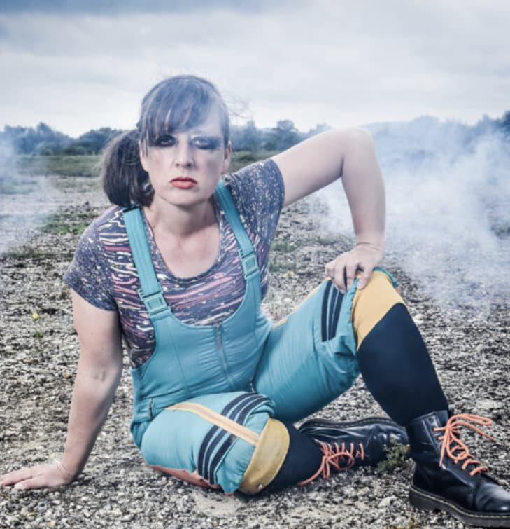 Image of a person in a green ski-suit sitting outdoors on a textured rocky surface with some smoke drifitng past.