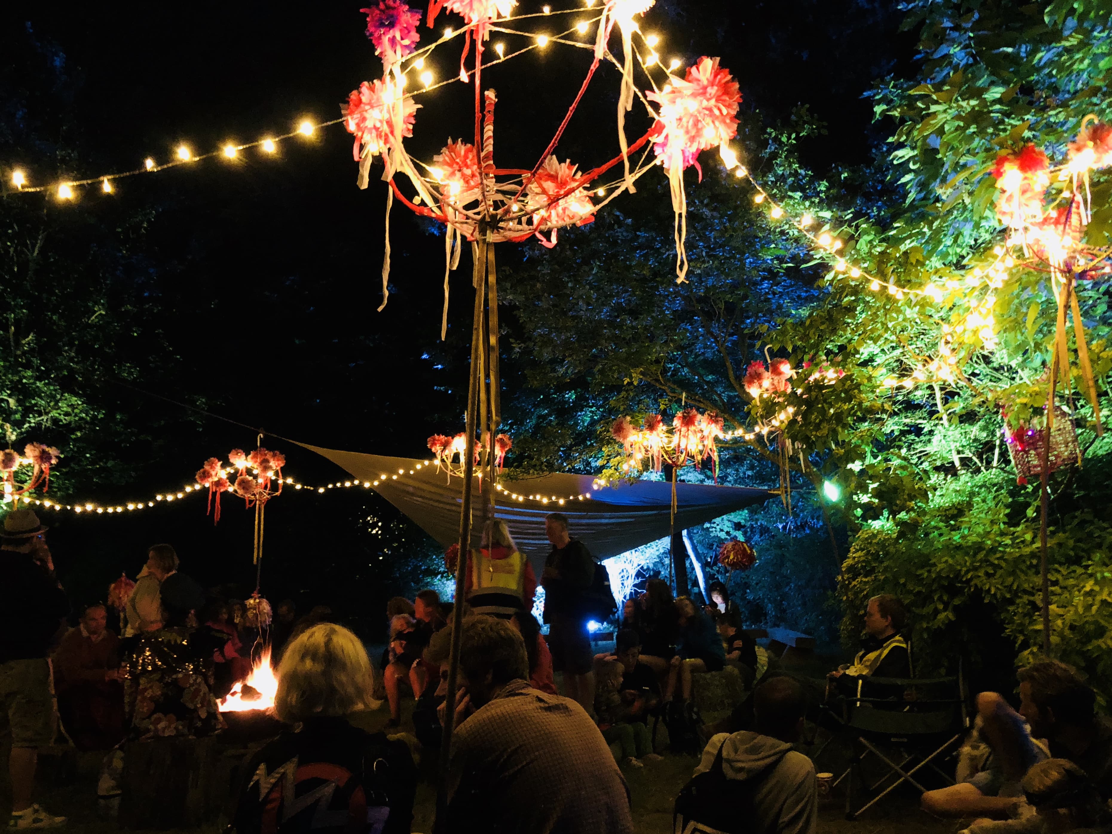 An image at night of a fesitval field with fairylight hung in a circle from stakes to create a space to sit and relax. Decorated with ribbons and colourful accessories.